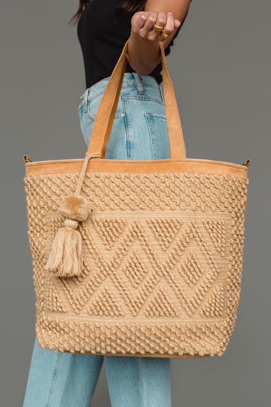 A person wearing a black shirt and blue jeans holds the panacheaccessories Tan Diamond Pattern Textured Tote, featuring a leather shoulder strap, against a plain gray background.