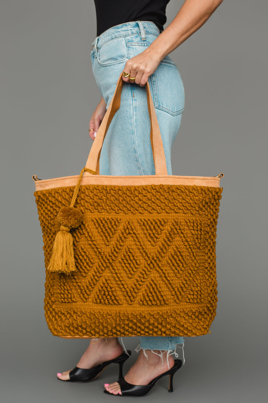 A person dressed in light blue jeans and black heels carries the Panache Apparel Collection Mustard Diamond Pattern Textured Tote, featuring a tassel detail and tan straps.