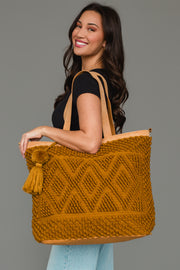 A woman with long dark hair smiles while holding the Mustard Diamond Pattern Textured Tote from Panache Apparel Collection, which features diamond patterns and a tassel. She wears a black top and light blue jeans, standing against a plain gray background.