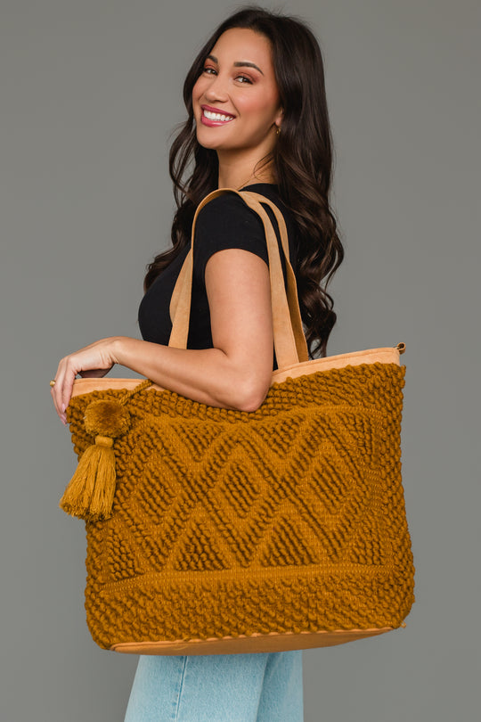 A woman with long dark hair smiles while holding the Mustard Diamond Pattern Textured Tote from Panache Apparel Collection, which features diamond patterns and a tassel. She wears a black top and light blue jeans, standing against a plain gray background.