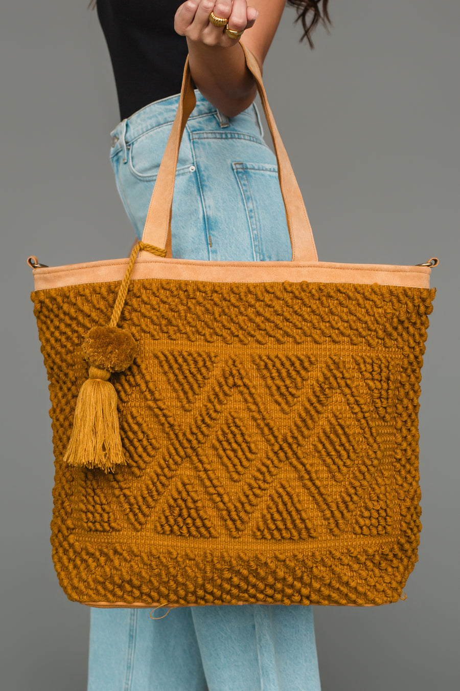A person in blue jeans and a black top holds the Panache Apparel Collection Mustard Diamond Pattern Textured Tote, featuring geometric patterns, leather straps, and a tassel accent.