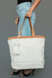 Wearing light blue jeans and black heels, a person holds the Panache Apparel Collection Ivory Diamond Pattern Textured Tote, featuring tan handles and a tassel detail, against a gray background.