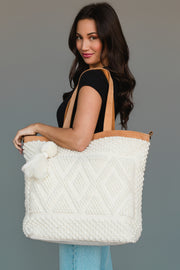 A woman with long dark hair holds the Panache Apparel Collection’s Ivory Diamond Pattern Textured Tote, featuring tan straps and a white tassel, while wearing a black top and light blue jeans against a plain gray background.