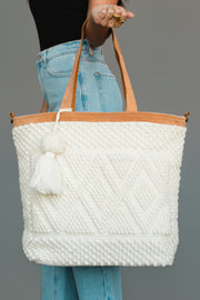 A person wearing blue jeans and a black top holds the Panache Apparel Collection Ivory Diamond Pattern Textured Tote, featuring brown handles and a white tassel, against a plain gray background.