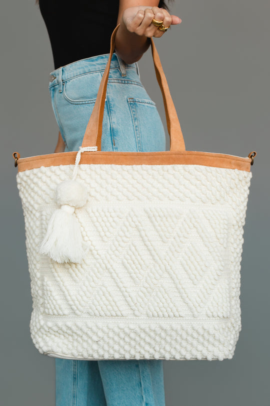 A person wearing blue jeans and a black top holds the Panache Apparel Collection Ivory Diamond Pattern Textured Tote, featuring brown handles and a white tassel, against a plain gray background.