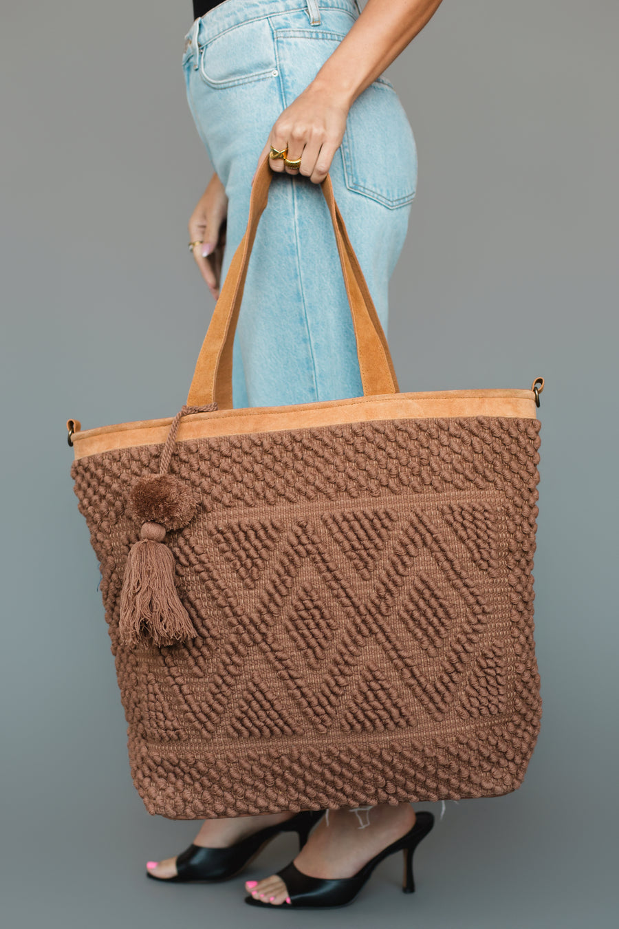 A person in light blue jeans and black heels holds the Brown Diamond Pattern Textured Tote by Panache Apparel Collection, featuring geometric patterns, leather trim, and a tassel, against a plain gray background.