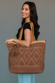 A woman with long dark hair and a black shirt smiles while holding the Panache Apparel Collection Brown Diamond Pattern Textured Tote, featuring a diamond pattern and tassel, against a plain gray background.