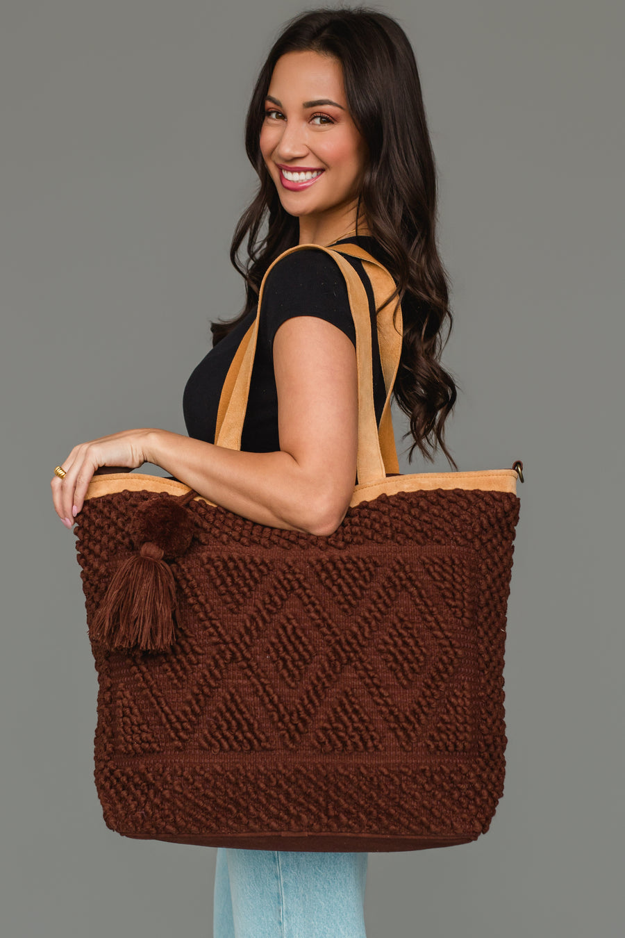 A woman in a black top and jeans smiles while holding the Panache Apparel Collection Dark Brown Diamond Pattern Textured Tote, featuring tan straps and a tassel, standing against a plain gray background.