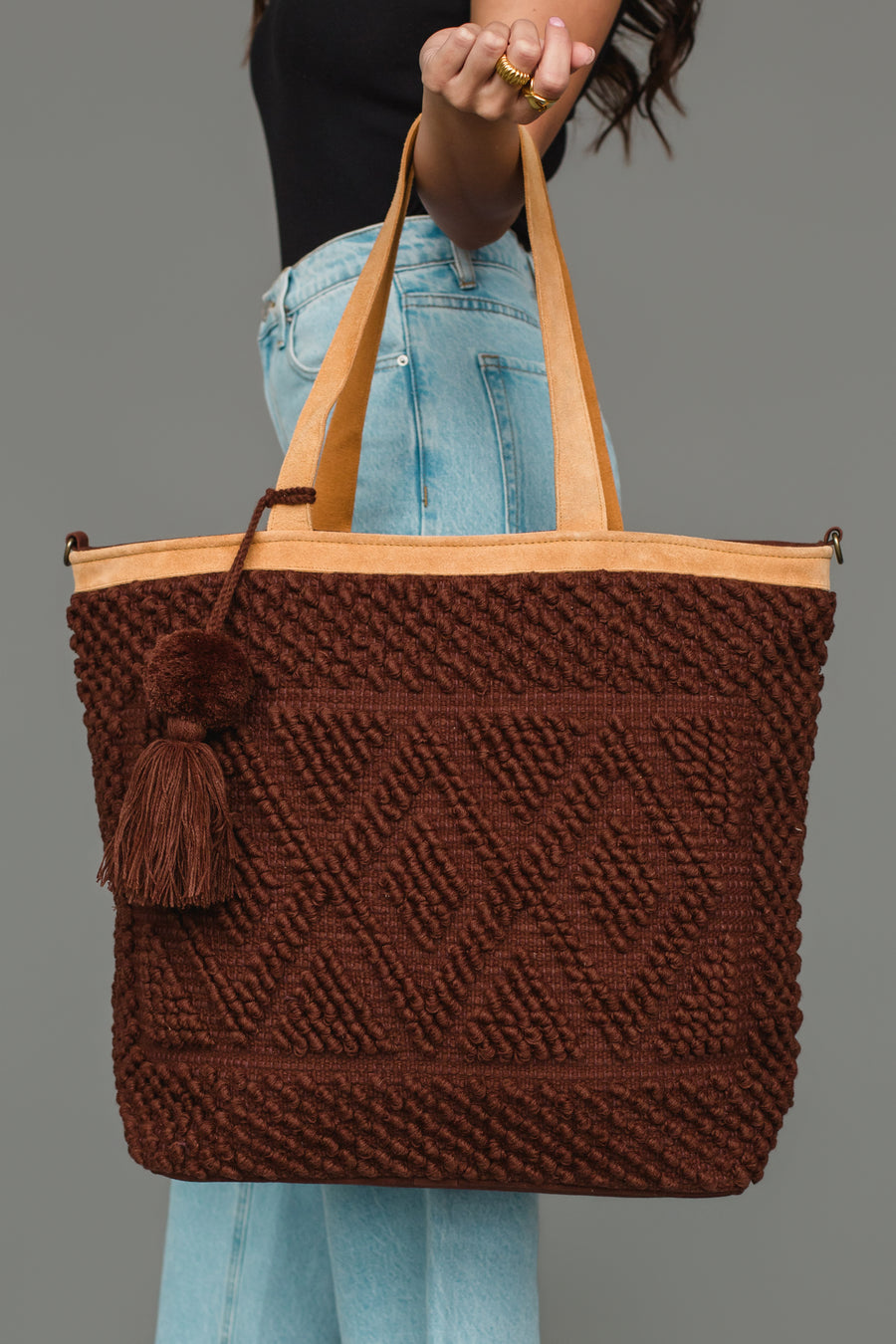 A person in jeans and a black top holds the Panache Apparel Collection’s Dark Brown Diamond Pattern Textured Tote, featuring leather handles and a tassel, against a plain gray background.