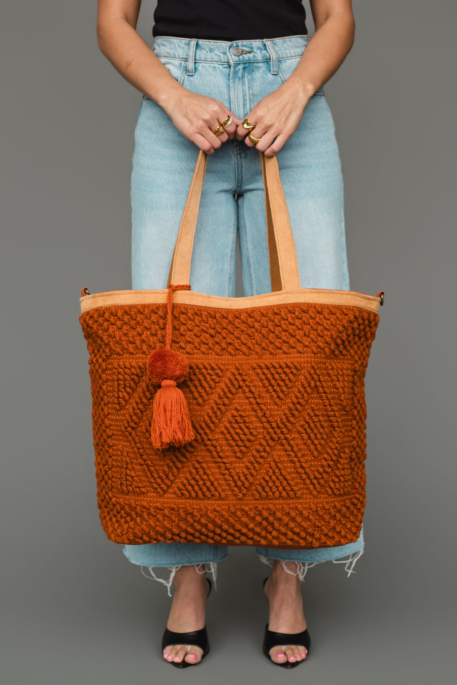A person in a black top and light blue jeans holds the Panache Apparel Collection Rust Diamond Pattern Textured Tote, featuring a tassel, while standing against a gray background.