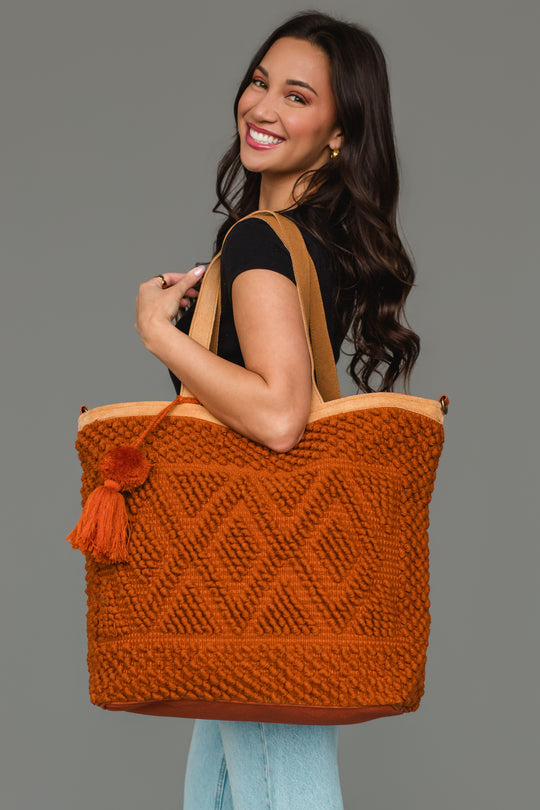 A woman smiles while wearing a black top and light jeans, holding the Panache Apparel Collection Rust Diamond Pattern Textured Tote, featuring a spacious design and tassel detail, against a plain gray background.