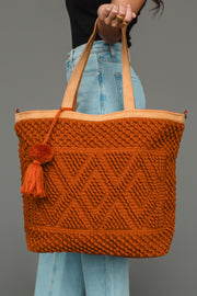A person in blue jeans holds the Rust Diamond Pattern Textured Tote from Panache Apparel Collection, its spacious design and geometric diamond pattern with a tassel showcasing a rich rust texture against the plain gray background.