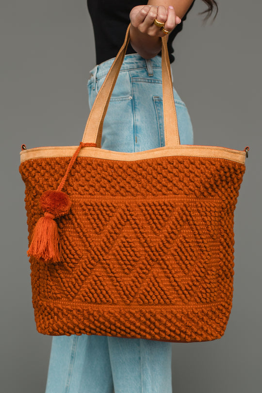 A person in blue jeans holds the Rust Diamond Pattern Textured Tote from Panache Apparel Collection, its spacious design and geometric diamond pattern with a tassel showcasing a rich rust texture against the plain gray background.