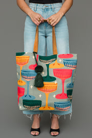 A person holds a Multicolored Coupe Glasses Tote featuring a pattern of coupe glasses and a dark green tassel.