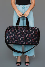 A person in light blue jeans and a black top holds the Panache Apparel Collection “Burgundy, Blue & Pink Floral Duffel” against a plain gray background, with only their lower legs and hands visible.