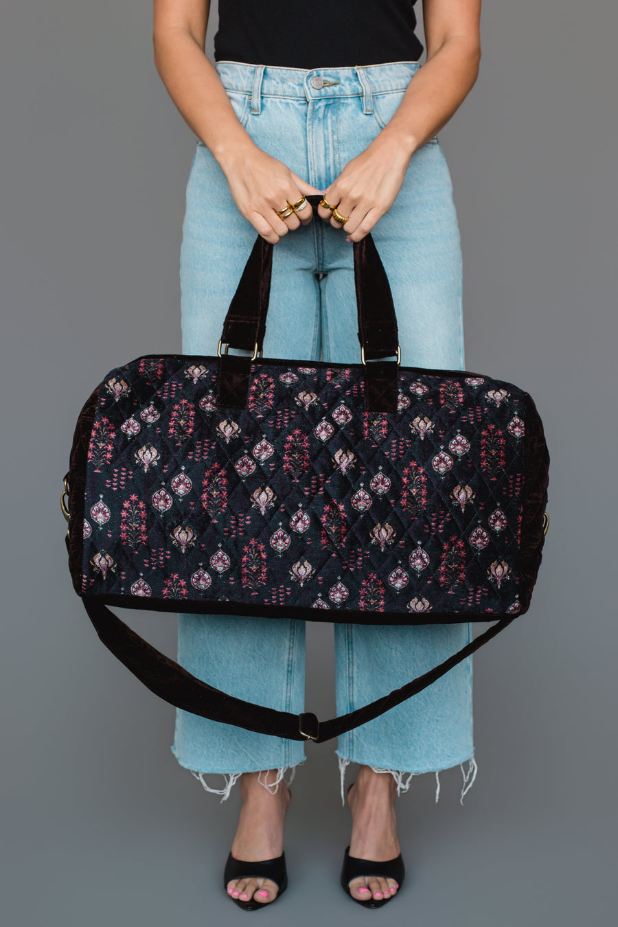 A person in light blue jeans and a black top holds the Panache Apparel Collection “Burgundy, Blue & Pink Floral Duffel” against a plain gray background, with only their lower legs and hands visible.