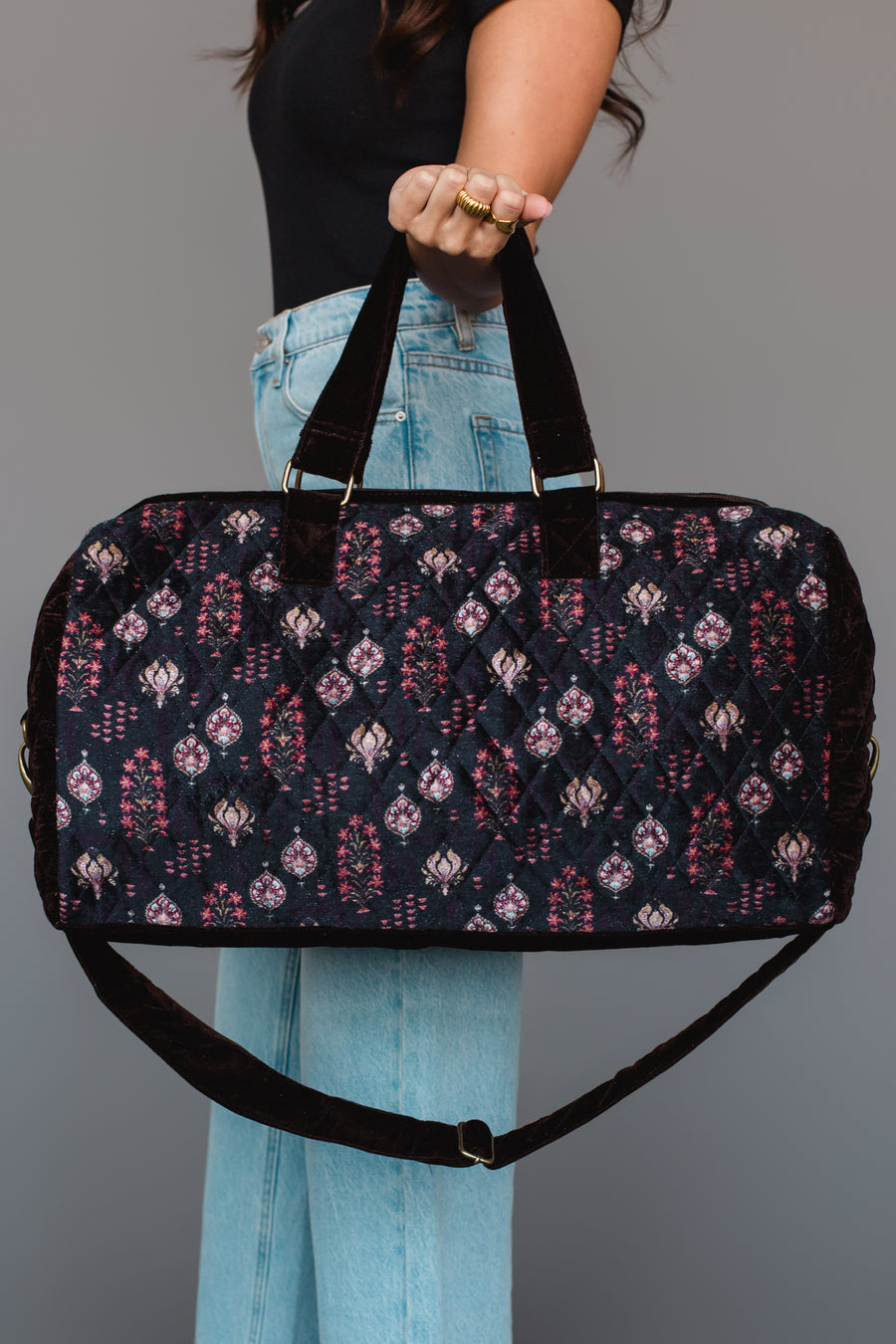 A person in a black top and light blue jeans holds the Panache Apparel Collection Burgundy, Blue & Pink Floral Duffel against a plain gray background.