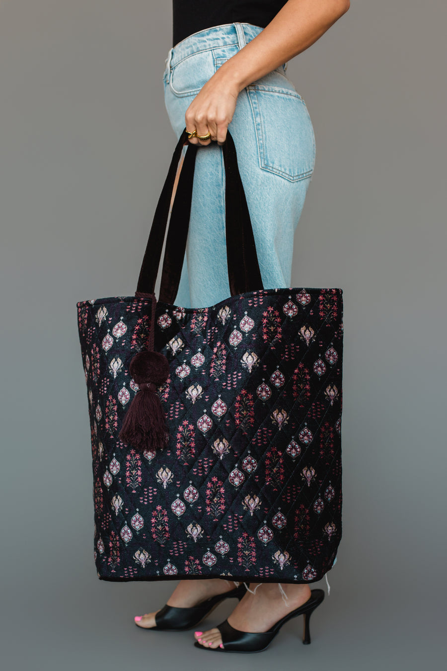 A person wearing blue jeans and black heels holds the Panache Apparel Collection Burgundy, Blue & Pink Floral Tote, a large, quilted black bag featuring a vibrant floral pattern, black straps, and tassel detailing, against a plain gray background.