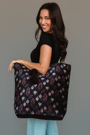 A woman with long dark hair, dressed in a black top and light blue jeans, smiles while holding the Panache Apparel Collection Burgundy, Blue & Pink Floral Tote, a large quilted bag featuring a floral pattern and pom-pom accents, over her shoulder against a plain background.