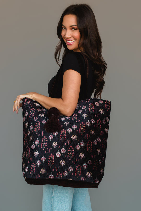 A woman with long dark hair, dressed in a black top and light blue jeans, smiles while holding the Panache Apparel Collection Burgundy, Blue & Pink Floral Tote, a large quilted bag featuring a floral pattern and pom-pom accents, over her shoulder against a plain background.