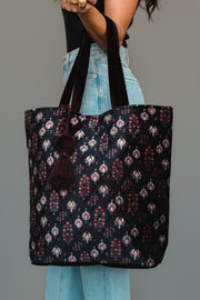 A woman holds the Panache Apparel Collection Burgundy, Blue & Pink Floral Tote, a large bag featuring colorful floral patterns and a dark tassel, while wearing a black sleeveless top and light blue jeans against a plain gray background.