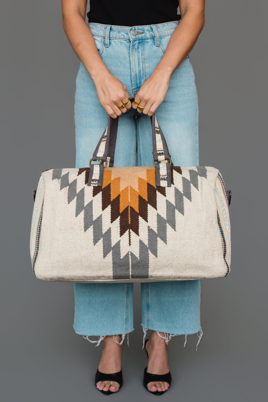 A person in light blue jeans and black shoes holds the Panache Apparel Collection’s White, Gray & Camel Western Duffel against a gray background.