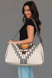 A woman smiles while wearing a black shirt and light blue jeans, holding the Panache Apparel Collection’s White, Gray & Camel Western Duffel, an oversized bag featuring a geometric tribal pattern in gray, orange, and brown with a patterned shoulder strap, set against a plain gray background.
