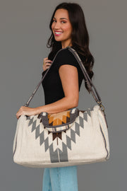 A woman with long dark hair, dressed in a black top and light blue jeans, smiles while holding the Panache Apparel Collection’s White, Gray & Camel Western Duffel against a plain gray background.