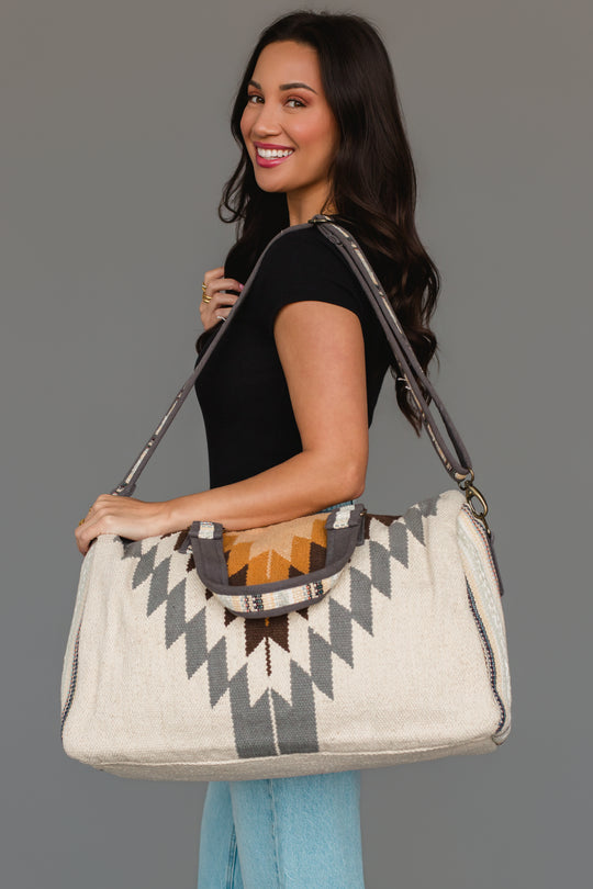 A woman with long dark hair, dressed in a black top and light blue jeans, smiles while holding the Panache Apparel Collection’s White, Gray & Camel Western Duffel against a plain gray background.