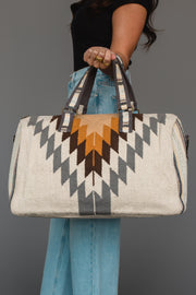 A person in jeans and a black top holds the Panache Apparel Collection White, Gray & Camel Western Duffel, featuring geometric patterns in white, gray, and camel tones. The bag is designed with dark handles and metal accents.