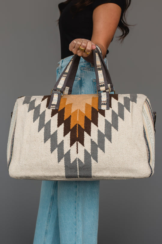 A person in jeans and a black top holds the Panache Apparel Collection White, Gray & Camel Western Duffel, featuring geometric patterns in white, gray, and camel tones. The bag is designed with dark handles and metal accents.