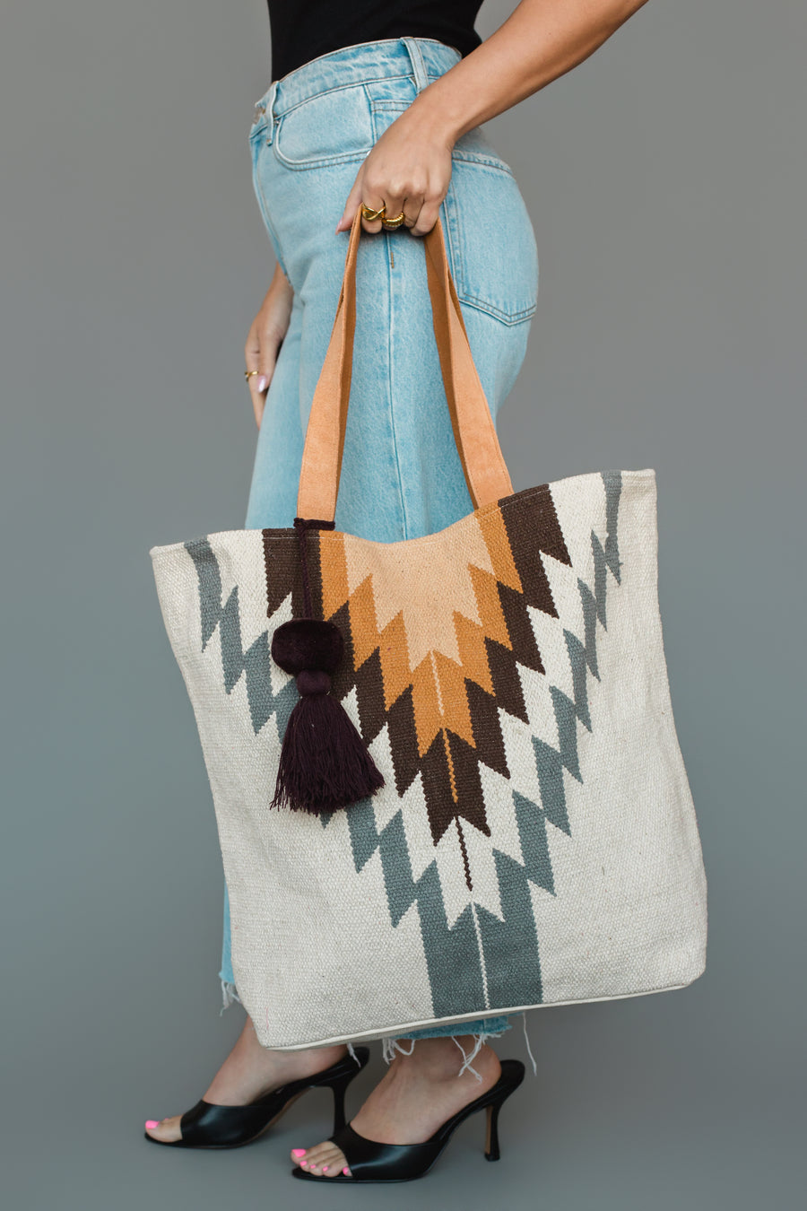 A person in jeans and heels holds the Panache Apparel Collection’s White, Gray & Camel Western Tote, a large woven bag with geometric patterns and leather handles, accented with a dark tassel.