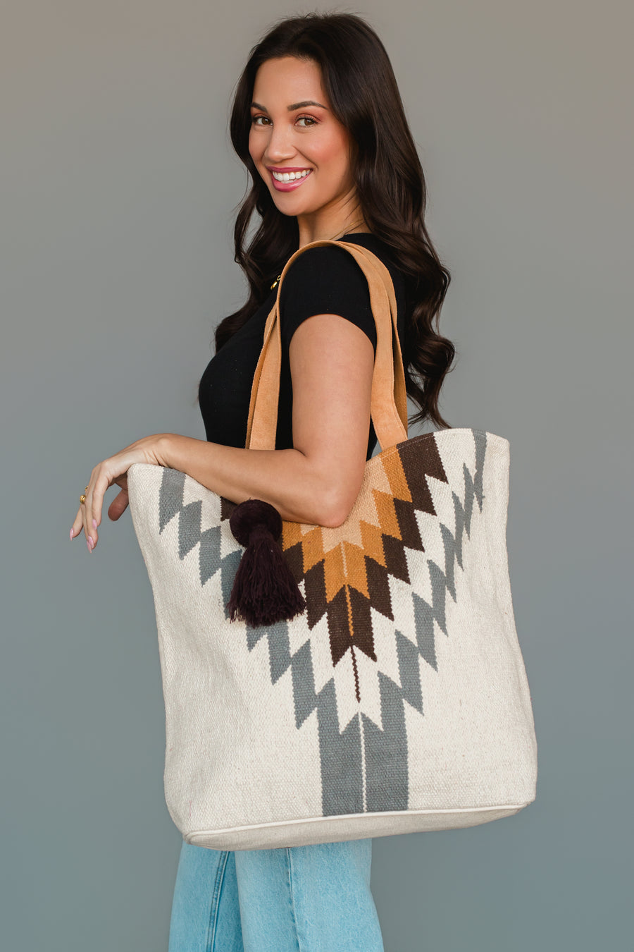 A woman smiles as she wears a black top and light jeans, holding the Panache Apparel Collection’s White, Gray & Camel Western Tote, a large cream bag featuring a geometric zigzag pattern in gray, brown, and tan with a dark tassel, against a plain gray background.