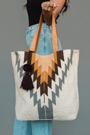 A person holding the Panache Apparel Collection White, Gray & Camel Western Tote, featuring zigzag patterns in brown, mustard, and gray with tan handles and a dark tassel, paired with light blue jeans and a black top.