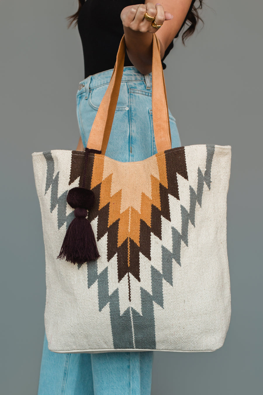 A person holding the Panache Apparel Collection White, Gray & Camel Western Tote, featuring zigzag patterns in brown, mustard, and gray with tan handles and a dark tassel, paired with light blue jeans and a black top.