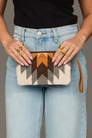 A person wearing light blue jeans and a black top holds the White, Gray & Camel Western Wristlet by Panache Apparel Collection, featuring a brown, gray, and beige geometric pattern. The person has manicured nails and wears multiple gold rings.