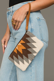 A person wearing light blue jeans holds the Panache Apparel Collection White, Gray & Camel Western Wristlet, which features a woven geometric zigzag pattern and a brown wrist strap.