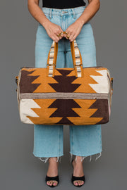 A person in blue jeans holds the Panache Apparel Collection “Camel, Brown & Cream Western Duffel,” featuring a geometric brown, orange, and cream pattern, while standing against a gray background.