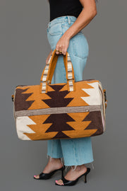 A woman in blue jeans and black heels holds the Camel, Brown & Cream Western Duffel from Panache Apparel Collection, featuring a bold geometric pattern in brown, tan, and cream. The duffel has two striped handles and a zip closure. The background is plain gray.