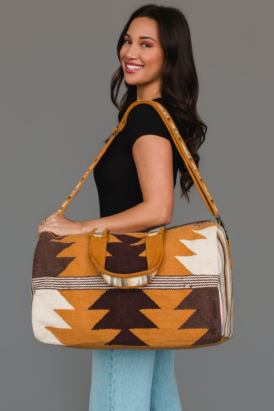 A woman with long dark hair, dressed in a black top and light blue jeans, smiles while carrying the Panache Apparel Collection Camel, Brown & Cream Western Duffel, featuring bold southwestern-inspired yellow, brown, and cream zigzag patterns, on her shoulder.