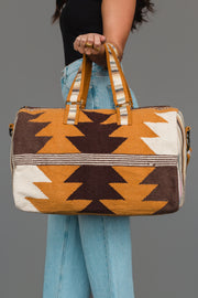 A person holds the Panache Apparel Collection Camel, Brown & Cream Western Duffel, featuring a geometric zigzag pattern in brown, mustard, and cream tones, while wearing blue jeans and a black top against a plain gray background.