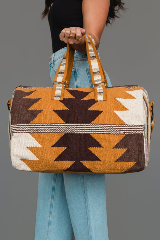 A person holds the Panache Apparel Collection Camel, Brown & Cream Western Duffel, featuring a geometric zigzag pattern in brown, mustard, and cream tones, while wearing blue jeans and a black top against a plain gray background.