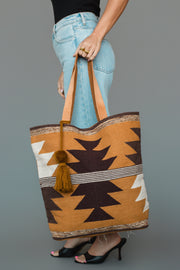A person in jeans and black heels holds the Panache Apparel Collection “Camel, Brown & Cream Western Tote,” a large woven bag featuring geometric brown, gold, and white patterns with a pom-pom tassel.