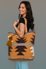 A smiling woman with long dark hair, dressed in a black top and light jeans, holds the Panache Apparel Collection Camel, Brown & Cream Western Tote, featuring a brown, mustard, and cream geometric pattern with pompom detail, while standing against a plain gray background.