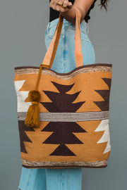 A person holds the Camel, Brown & Cream Western Tote by Panache Apparel Collection, featuring tan, brown, and cream geometric patterns with a tassel. They are wearing light blue jeans and a black top.