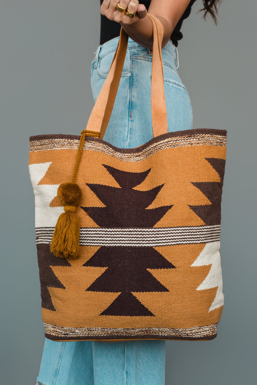 A person holds the Camel, Brown & Cream Western Tote by Panache Apparel Collection, featuring tan, brown, and cream geometric patterns with a tassel. They are wearing light blue jeans and a black top.