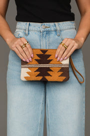 A person in light blue jeans and a black top holds the Panache Apparel Collection Camel, Brown & Cream Western Wristlet, a small woven clutch featuring a geometric pattern in brown, orange, and cream hues. Their nails are light pink and they are wearing multiple gold rings.