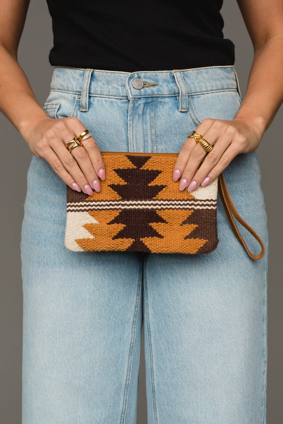 A person in light blue jeans and a black top holds the Panache Apparel Collection Camel, Brown & Cream Western Wristlet, a small woven clutch featuring a geometric pattern in brown, orange, and cream hues. Their nails are light pink and they are wearing multiple gold rings.