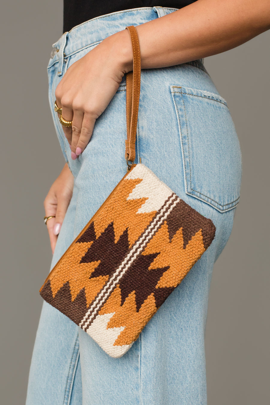 A person wearing light blue jeans holds the Panache Apparel Collection Camel, Brown & Cream Western Wristlet, which features a brown, orange, and cream geometric pattern and a tan wrist strap.