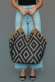 A person in light blue jeans holds the Panache Apparel Collection Black & Cream Diamond Pattern Textured Tote, featuring brown handles. Only their lower torso and legs are visible against a plain gray background.
